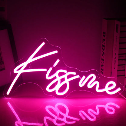 A vibrant "Kiss Me" LED neon sign is illuminated, displaying bright and bold pink lettering against a dark background.
