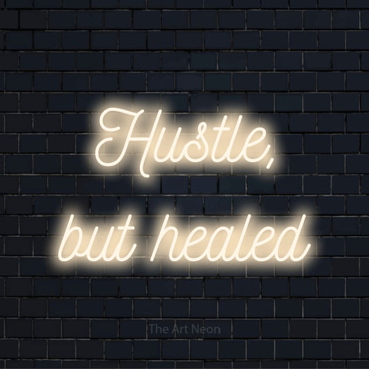 "Hustle, But Healed" script-style LED neon sign on a black brick wall