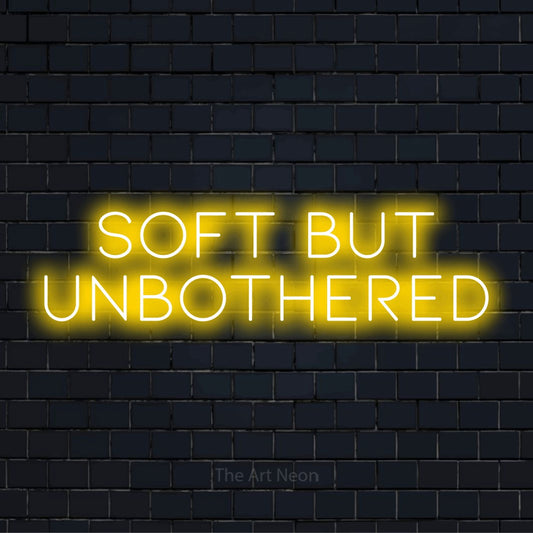 "Soft But Unbothered" script-style LED neon sign on a black brick wall