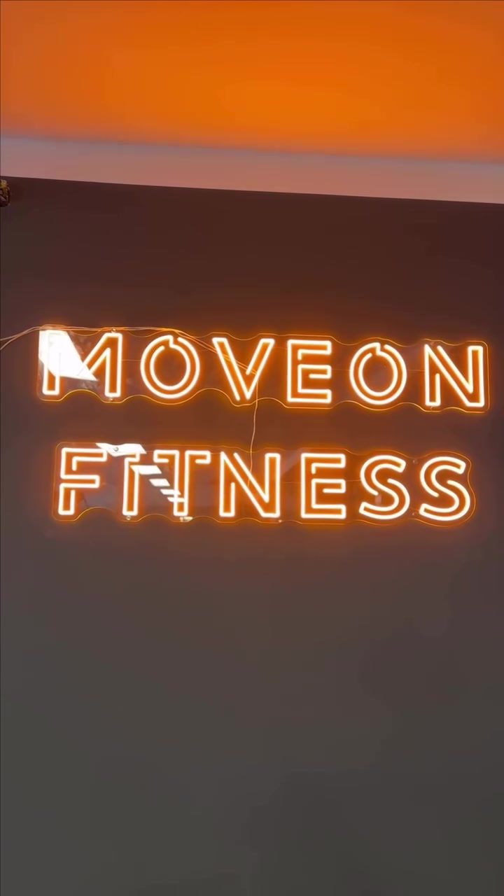 Warm-yellow MOVEON FITNESS LED neon logo mounted on a dark fitness studio wall