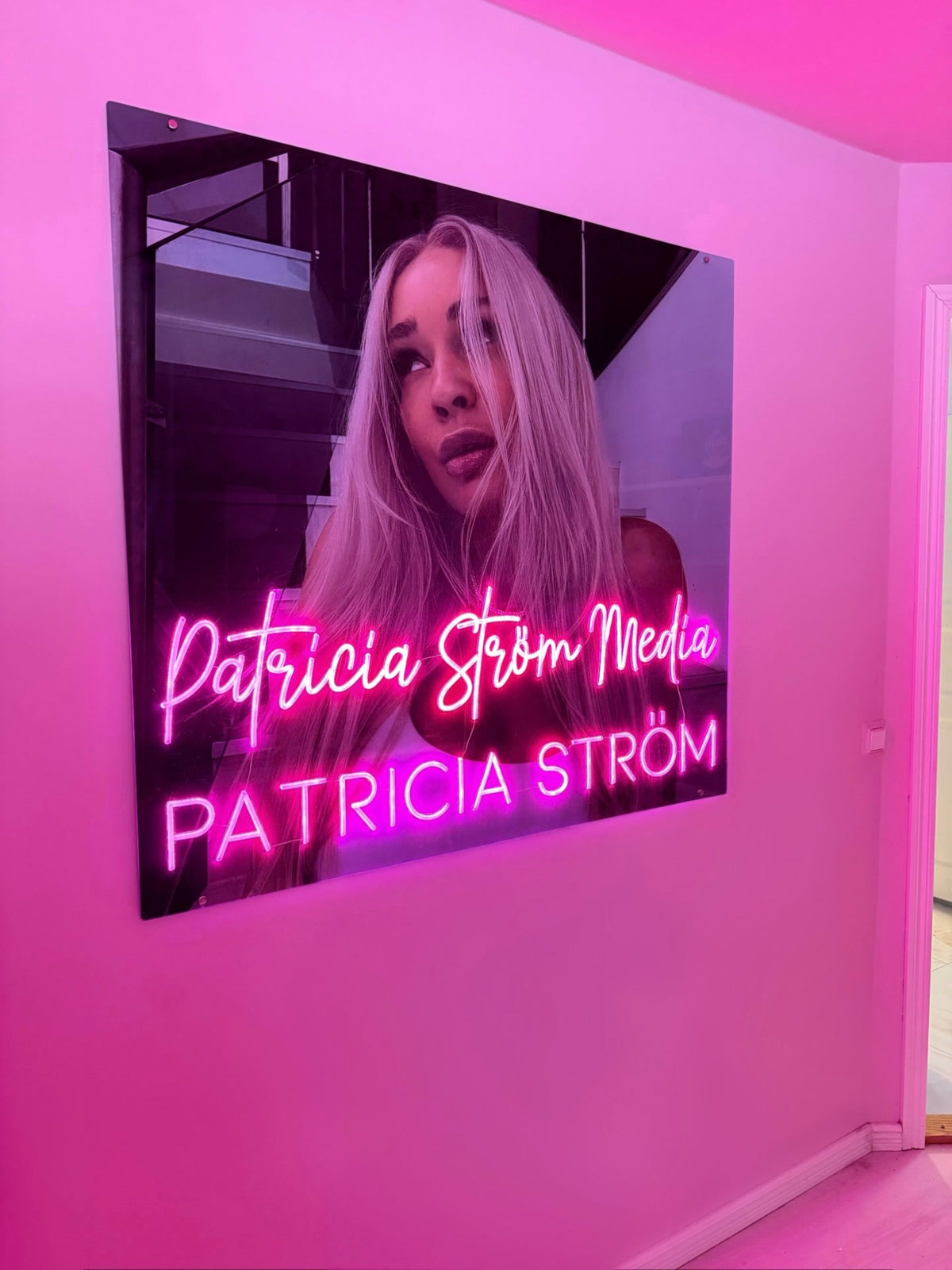 Roze Patricia Ström Media-neonlogo naast Patricia Ström in haar werkruimte met een persoonlijke merkuitstraling
