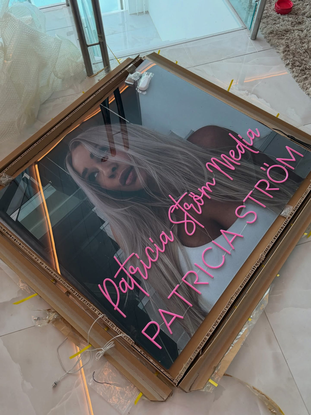 Roze Patricia Ström Media-neonlogo naast Patricia Ström in haar werkruimte met een persoonlijke merkuitstraling
