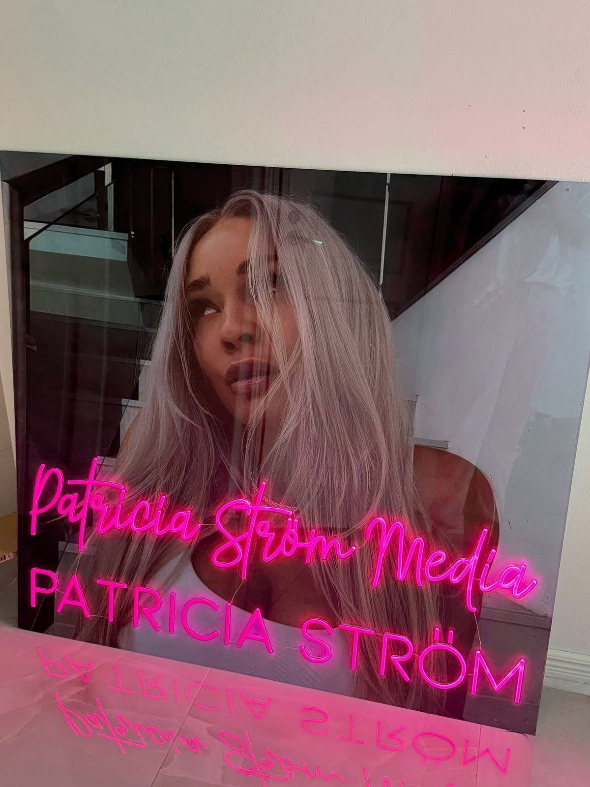 Roze Patricia Ström Media-neonlogo naast Patricia Ström in haar werkruimte met een persoonlijke merkuitstraling